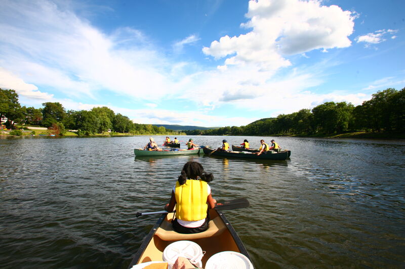 The image shows a sunny day on a lake with several canoes. In the foreground, a person is sitting in a canoe, facing away from the viewer, with a paddle resting in the boat. Further out, two canoes are filled with people paddling across the water. The background features lush green trees lining the shore under a bright, partly cloudy sky.